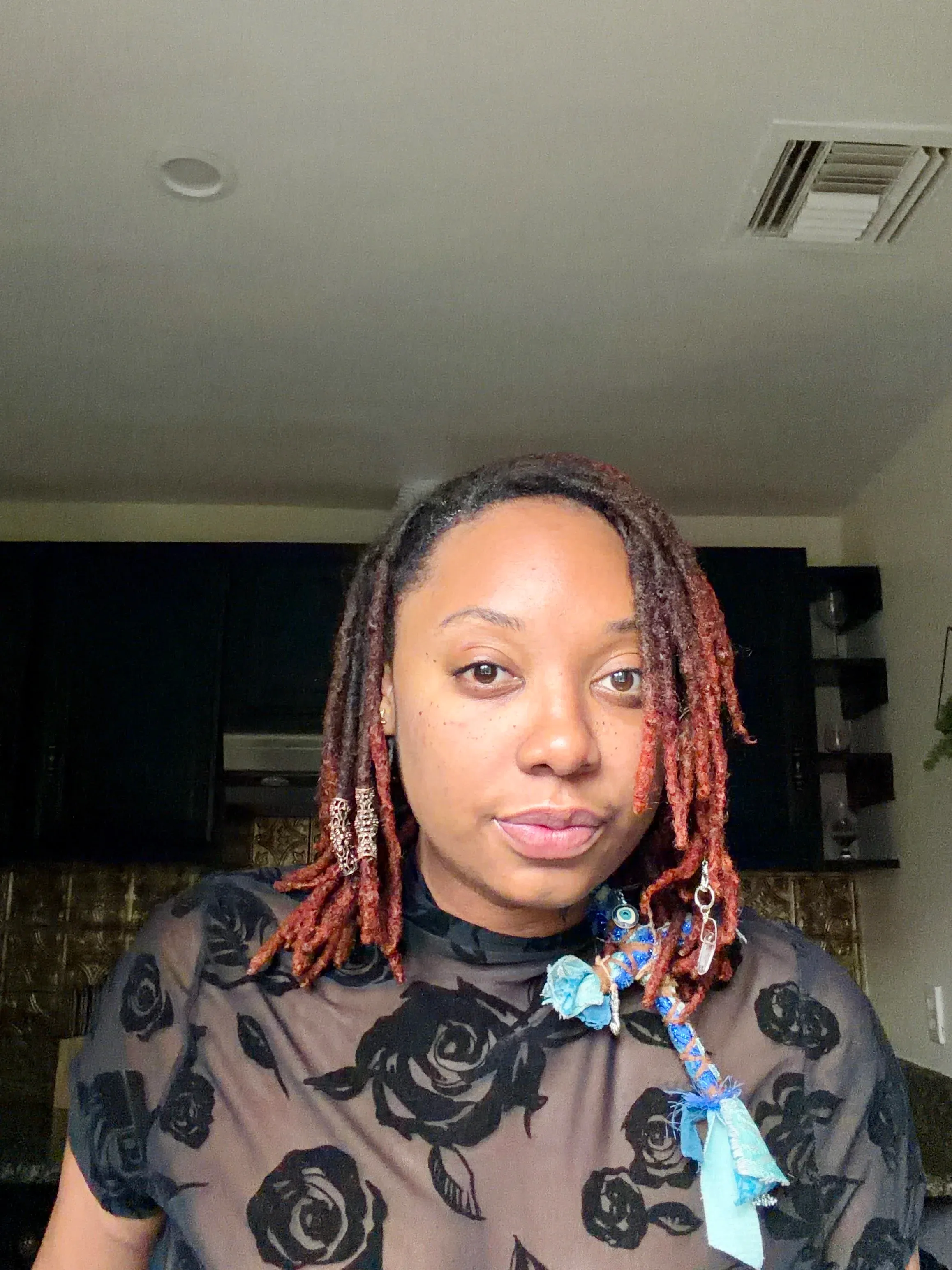 Becca Berry looking directly at the camera, with russet locs and a sheer black rose-print top.
