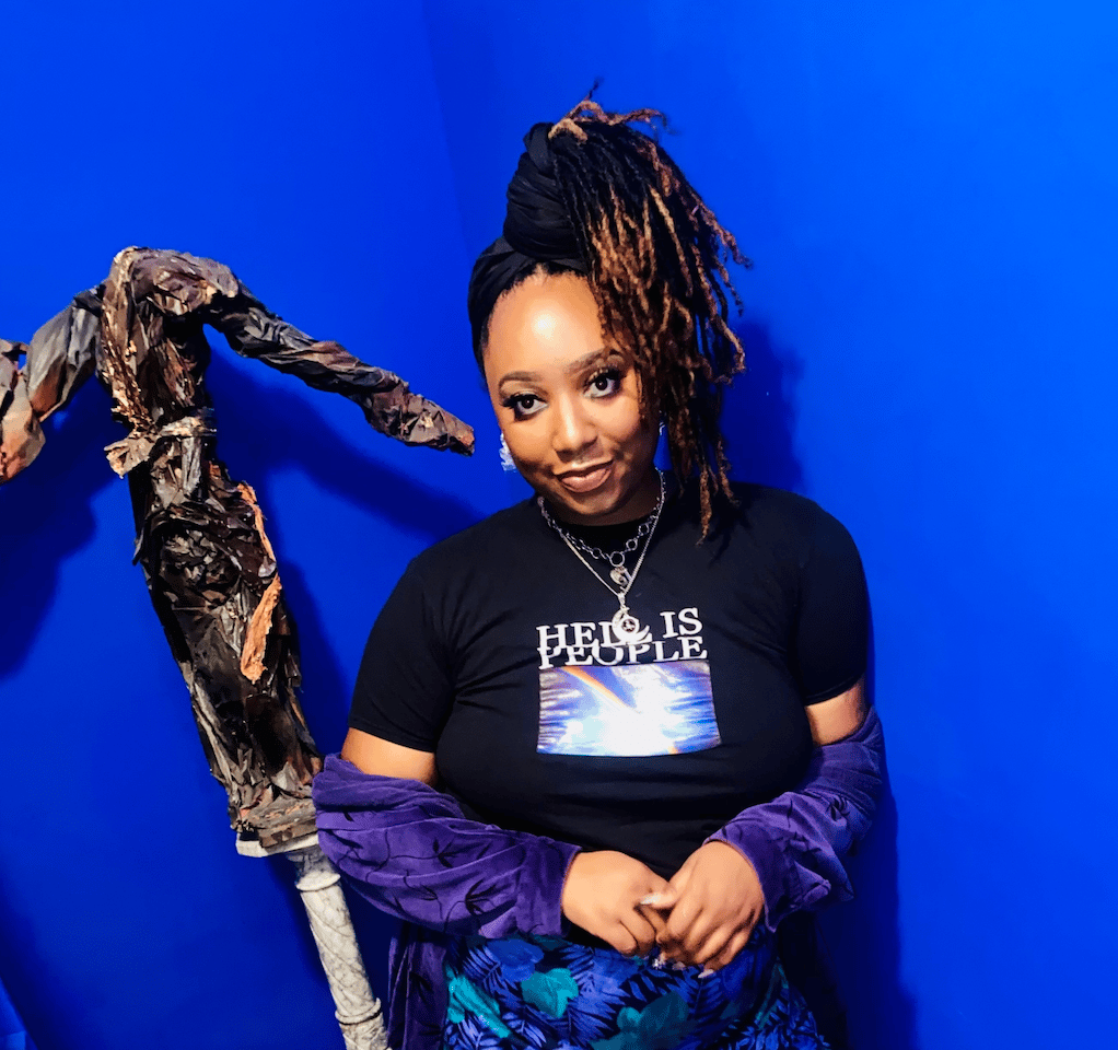 Becca Berry looking directly at the camera, with russet locs and a sheer black rose-print top.